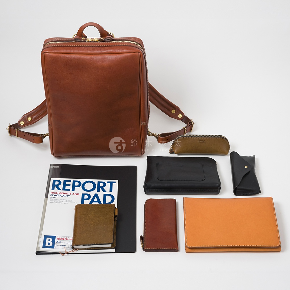 一個棕色皮革商務後背包，旁邊擺放著一個綠棕色鉛筆盒、一個黑色長夾、一個黑色卡夾、一個棕色筆記本、一個棕色皮夾和一個橘色文件夾。