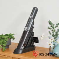 充電式手持吸塵器，深灰色機身，搭配黑色充電座，放置於木質檯面上，左側有綠植，右側有花瓶。