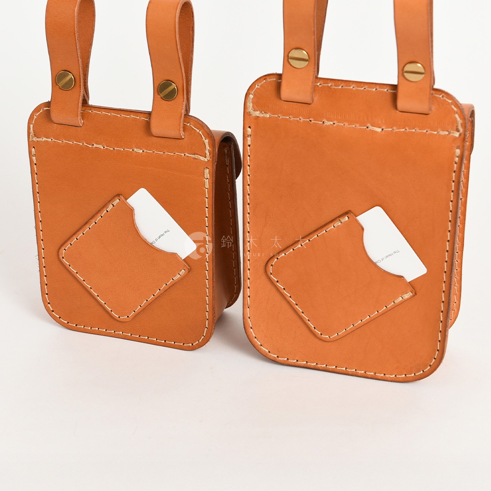 兩款職人手作皮革經典腰袋，皆為棕褐色，帶有明顯縫線。前方均有一個菱形小口袋，各插有一張白色卡片。腰袋上部有兩條皮革掛帶，以金屬釦固定。