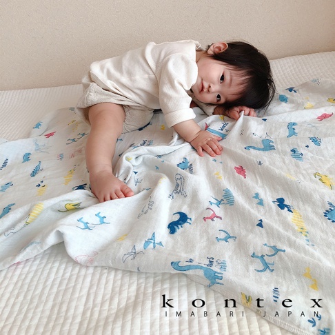 一嬰兒趴蓋在印有可愛動物塗鴉圖案的白色二層紗嬰兒毯上。毯子上有藍色、粉色和黃色的恐龍、鳥類和長頸鹿等圖案。