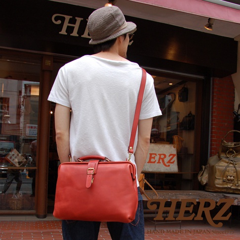 一位男士身穿白色T恤，肩背一個紅色軟式皮革杜勒斯包。包包為硬挺的長方形，頂部有提把，正面有一條皮帶束口，搭配金色扣環。