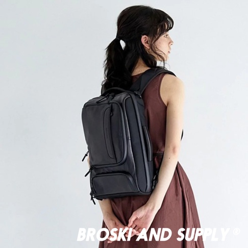 一位女性身著咖啡色無袖連身裙，背著一個黑色兩用手提後背包。背包採用防水真皮革材質，設計簡潔，有多個拉鍊隔層，包括正面一個較小的拉鍊口袋，以及兩個較大的主隔層。