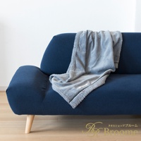 淺灰色羊毛感膝蓋毯，搭配深藍色沙發，毯子邊緣有毛絨細節，角落處有小標籤。