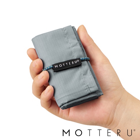手持一個摺疊起來的淺灰色隨行袋，袋子由輕薄的尼龍材質製成，並用藍色和黑色的彈性繩綁住，上面有一個寫有「MOTTERU」的黑色標籤。