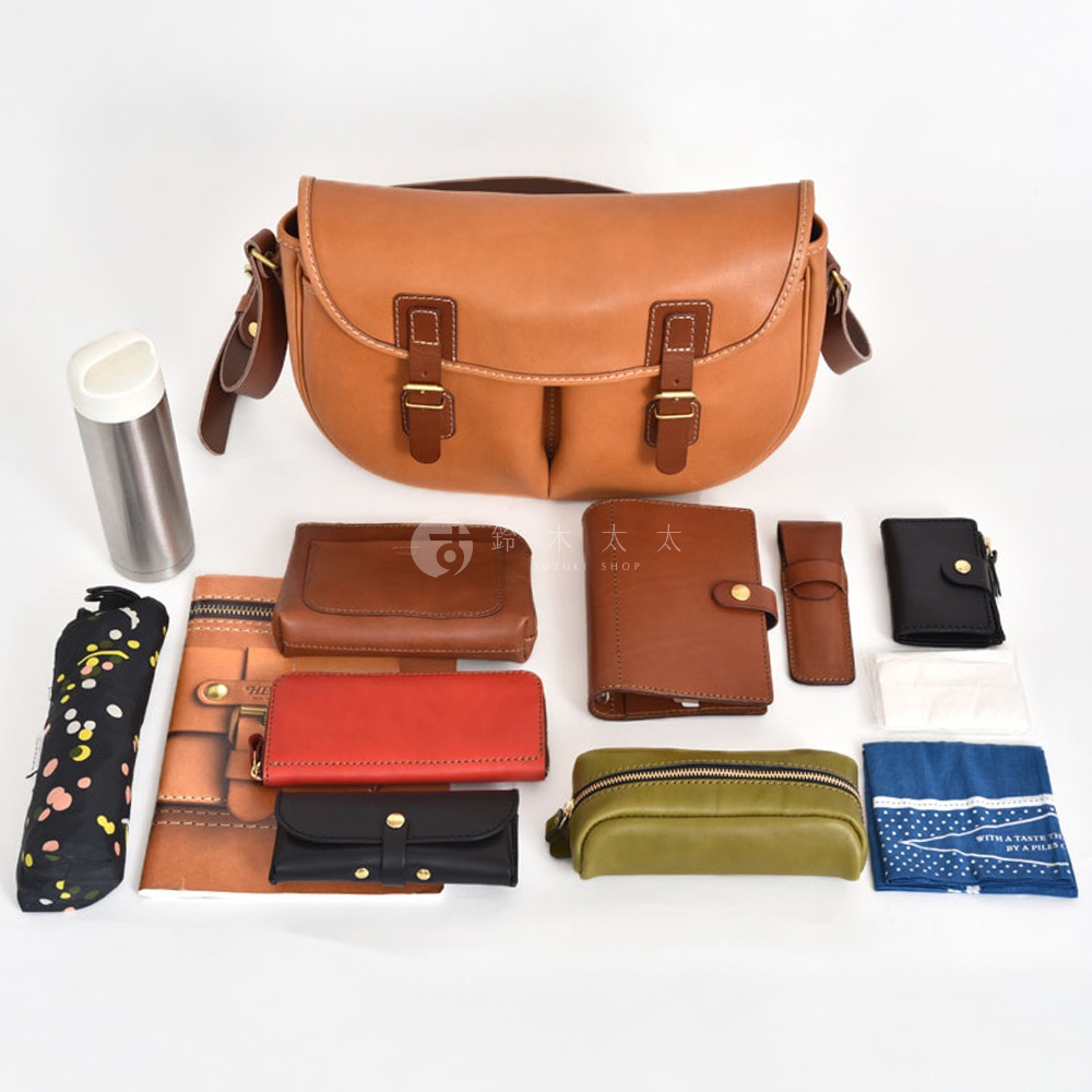 職人手作皮革傳統釣魚包，淺棕色皮革，附兩個皮帶扣。周圍展示多個配件，包括一個鋼製保溫瓶、一個黑色碎花雨傘、多個皮夾（棕色、紅色、黑色），以及一個綠色化妝包。