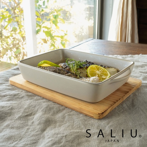 一個米白色長方形的 SALIU 耐熱陶製烤盤，放置在木質砧板上，烤盤內盛裝著煎魚料理，配有檸檬切片與新鮮香草點綴，背景為明亮的窗邊餐桌，展現出簡約自然的居家料理氛圍。