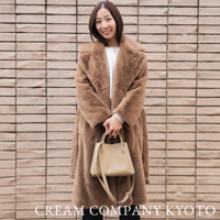 身穿棕色長款毛絨外套的女性，手持米色皮革兩用手提托特包，包款簡潔，搭配可拆卸肩帶。