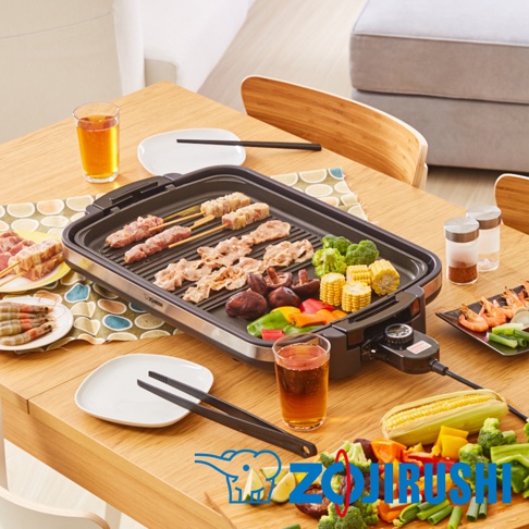 黑色分離式電烤盤，上面烤著串燒、肉片、香菇、玉米、青椒、紅椒和花椰菜。一旁有蝦仁、兩杯飲料，以及烤盤旁的黑色夾子和兩個調味罐。