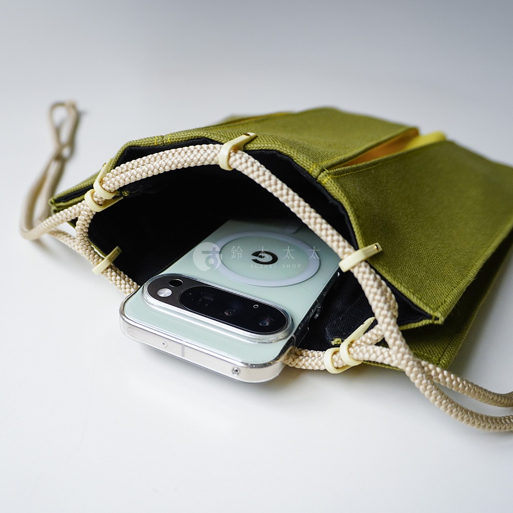 一個橄欖綠色的帆布信玄袋，袋口以米白色的繩索編織而成，繩索透過米白色塑料固定扣穿過袋緣。袋子內部鋪有黑色內裡，正放入一支背部帶有磁吸環的淺綠色智慧型手機，視角為由上往下俯拍，展現袋子的開口設計與質感。