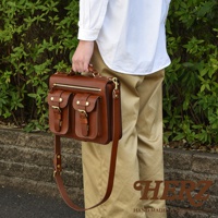 棕色皮革雙口袋兩用單肩包，配有金色拉鍊和搭扣。包款設有兩個前置翻蓋口袋，並可透過頂部手柄或可調式肩帶攜帶。