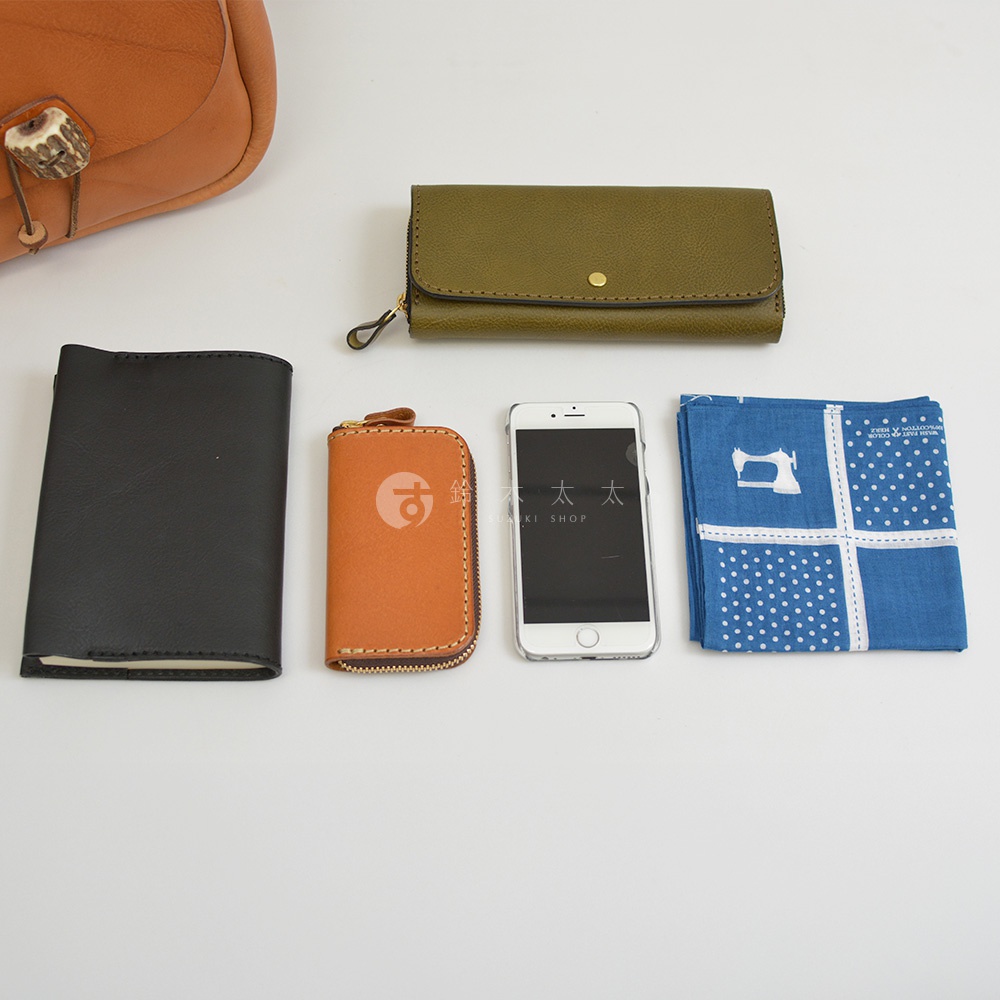 棕褐色皮革鹿角扣繩肩背包，旁邊擺放著黑色筆記本、橘色鑰匙包、白色手機及藍色印花手帕。