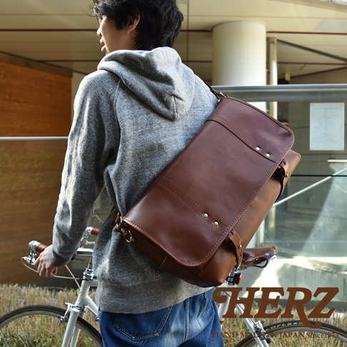 職人手作皮革復古郵差包，採用光滑的棕色皮革製成，配有金屬扣環和縫線細節，斜挎於身著灰色連帽衫和牛仔褲的男士肩上，整體展現復古風格。