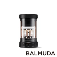 The BALMUDA wireless speaker is cylindrical in shape with a black top and base. A clear glass cylinder encloses three glowing vacuum tubes with copper accents. The base features control buttons for po