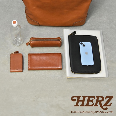 HERZ 職人手作的皮革配件，包含一個棕色托特包、一個長夾、一個筆袋、一個小零錢包，以及一個黑色收納包，包內放著一支手機。