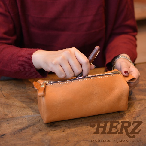 一個棕色皮革製成的職人手作多用途收納筆袋，拉鍊開口，一名女子正在將筆放入筆袋中。