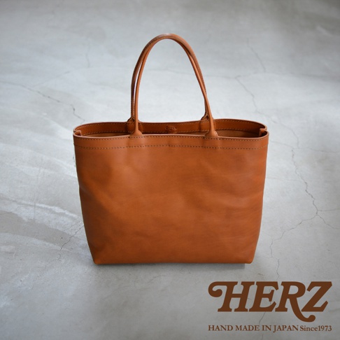 一個淺棕色皮革托特包，有兩個提把，包包底部和側面有縫線細節，背景是水泥紋理的地面。包的右下角有HERZ品牌標誌，下方有HAND MADE IN JAPAN Since1973字樣。