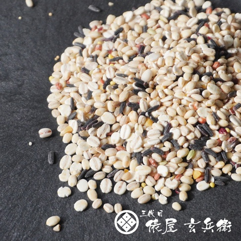 堆疊在黑色粗糙底板上的十六穀米，包含白色大麥、黑色米粒以及多種淺色雜糧，整體色澤豐富自然。右下角標示有白色圓形徽章圖樣與三代目俵屋玄兵衛的品牌中文字樣，展現出高品質日本進口穀物的原始風貌。