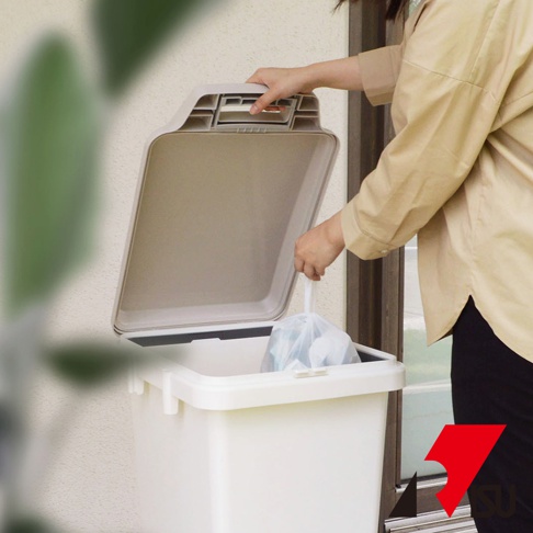 一個人正將垃圾袋放入一個米白色、掀蓋式的大容量垃圾桶。垃圾桶蓋為淺褐色，有防臭設計。