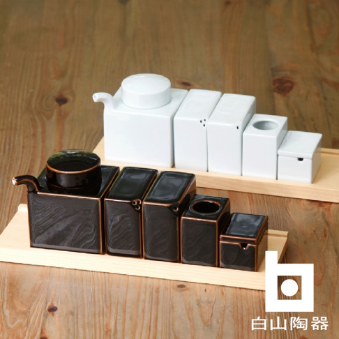 木盤上擺放著兩組陶瓷調味罐，左邊為黑色，帶有金屬光澤的邊緣，包含一個帶壺嘴的方形容器，上面放著一個圓形蓋子，以及四個不同大小的方形容器；右邊為白色，包含一個帶壺嘴的方形容器，上面放著一個圓形蓋子，以及四個不同大小的方形容器。