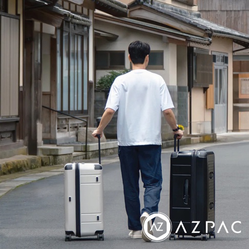 一名男子走在日式建築街道上，拉著一個銀色和一個黑色的 AZPAC 26 吋旅行箱，兩款皆為硬殼材質，配有防爆拉鍊和伸縮拉桿。