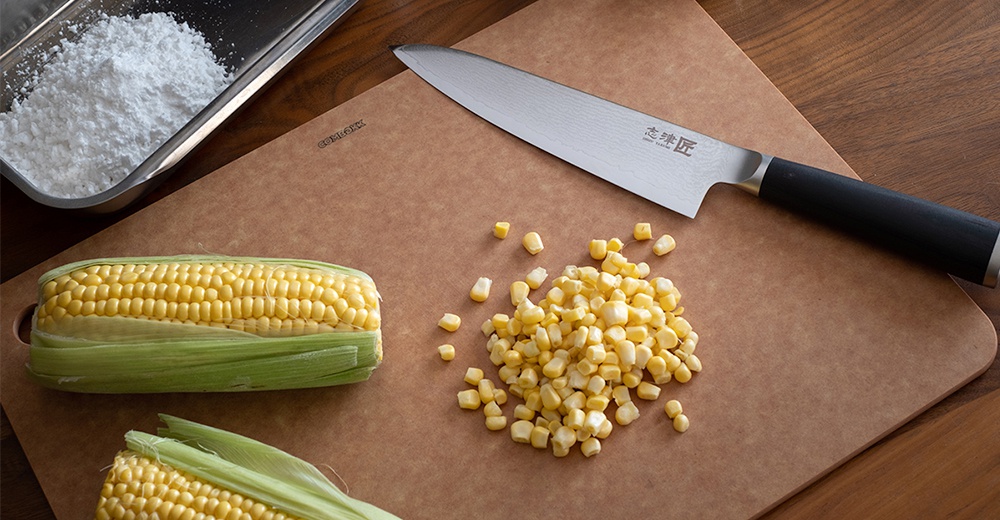 志津匠都水果刀旁有兩根玉米，其中一根已去皮，部分玉米粒被切下堆成一小堆，旁邊還有一小盆白色粉末。