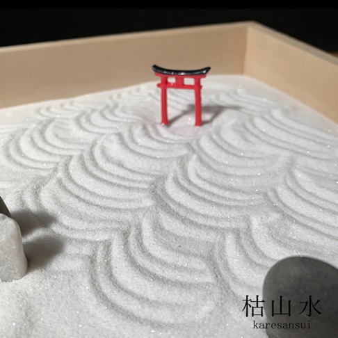 迷你枯山水模型，白色細沙上耙出波浪紋，中央立著紅色鳥居，左側有白色石塊，右下角有深色圓石及日文「枯山水」字樣。