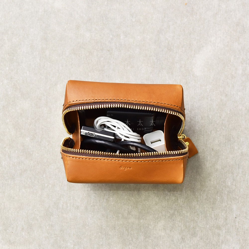 一個打開的棕褐色皮革箱型收納包，裡面裝有充電線、USB 充電器和一個黑色的小裝置。