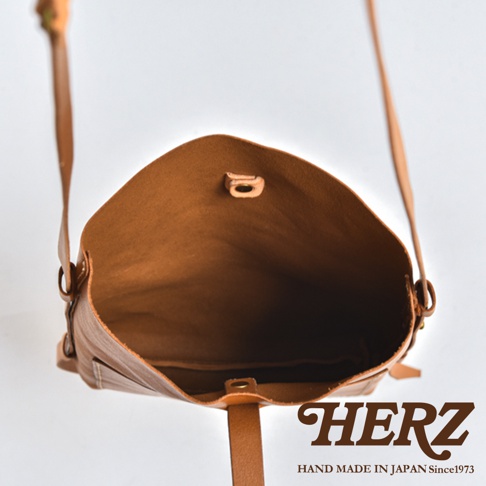 這張照片呈現日本職人品牌HERZ手作皮革單肩包的內部俯視視角，展示其簡潔俐落的包口結構與金屬釦環細節，整體採用溫潤的淺棕色皮革製作，右下角印有HERZ品牌商標與日本手工製作的相關說明文字。
