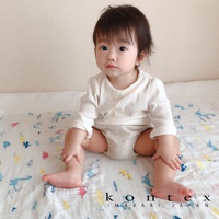 一名嬰兒身穿白色長袖上衣與米色尿布，坐在印有恐龍、兔子等童趣塗鴉圖案的二層紗嬰兒毯上。