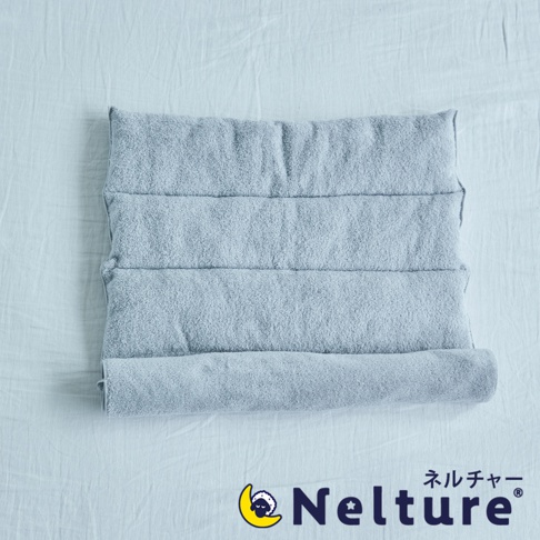 淺藍色毛巾材質的Nelture捲捲快眠今治毛巾枕，表面有四道縫線，一端捲起，放置於淺色紋理布料上。