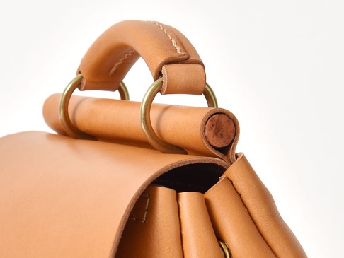 淺棕色皮革製作的迷你後背包，頂部有圓形提把，以黃銅色金屬環固定，皮革縫線清晰可見。