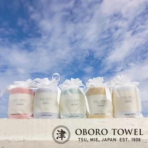 五條「潤澤素肌微笑速吸浴巾」以淡粉、淡藍、淡綠、米黃及白色五種柔和色彩呈現，皆以白色半透明包裝袋束口綁帶包裝，放置於白色石牆上，背景為藍天白雲。