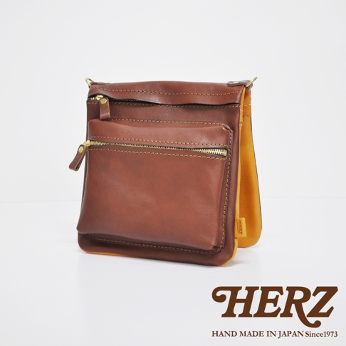 職人手作皮革翻轉雙色直式斜背包，主體為棕色，翻蓋內側為芥末黃色。包包採用拉鍊開合，正面有拉鍊口袋，皮革質感，縫線明顯。