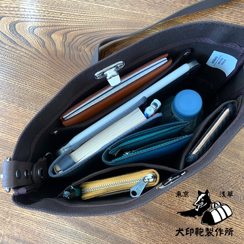 犬印純綿帆布雙色迷你側背包，深棕色帆布材質，包內裝有皮夾、筆記本、筆、手機、水瓶及黃色零錢包，整體呈現實用收納的畫面。