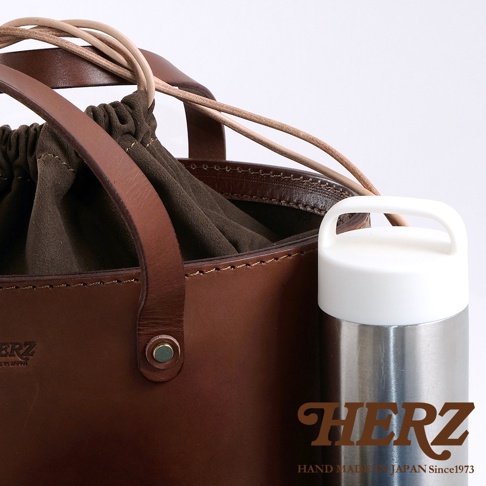 職人手作皮革束口手提包，採用棕色皮革製作，內有深棕色布料束口，搭配兩條淺棕色皮革提把，側面有一個金屬扣。