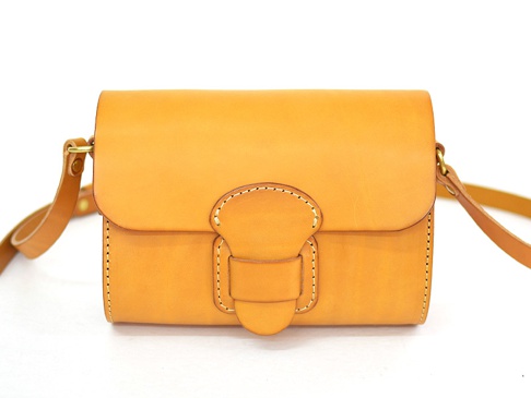 職人手作皮革寬扣帶馬鞍包，採用天然棕色皮革製成，包身結構方正，帶有精緻的縫線細節和寬版皮革扣帶，側邊有金屬掛環連接可調節的皮革背帶。