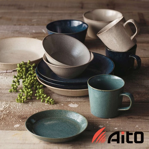 一組藍色、米色和灰色的陶製碗盤，包含盤子、碗和馬克杯，擺放在木製桌面上，旁邊有一叢綠色植物。