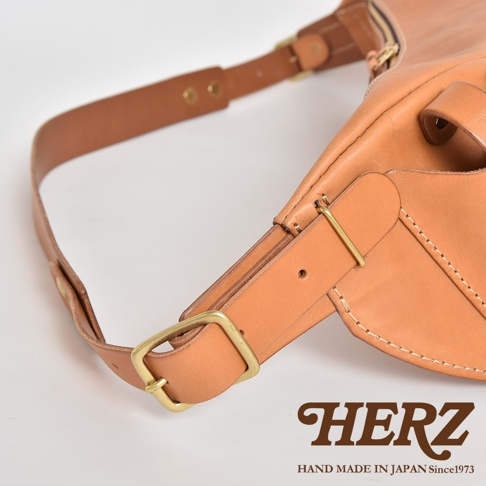 HERZ 品牌標誌和 HAND MADE IN JAPAN Since1973 字樣，旁邊為一個 tan 色皮革斜背包的細節，包含金屬扣環和皮革帶。