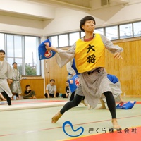 一位身穿和服、黃色背心的男子，手持藍色印有白色圖案的小抱枕，正準備投擲。背景有其他穿著類似服飾的人員和觀眾。