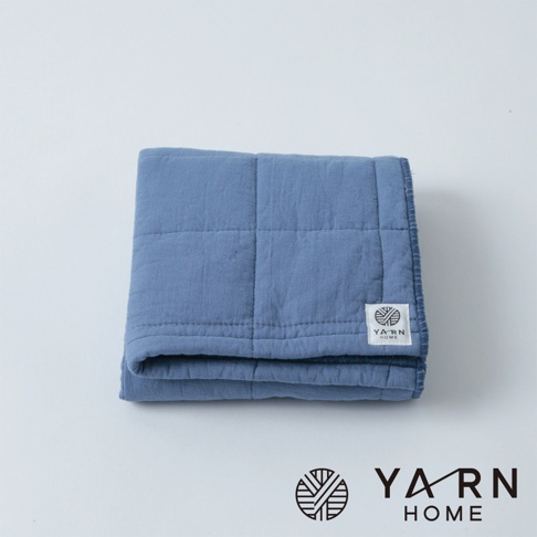 藍色格紋鋪棉毛巾，材質觸感柔軟，邊緣有細緻包邊，角落處有白色品牌標籤。