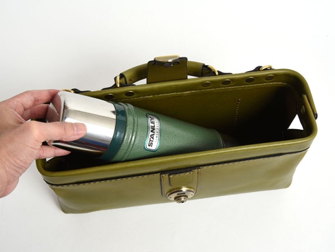 橄欖綠皮革製成的橫長形單肩包，包內放入一個綠色金屬保溫瓶，手正將其放入包中。