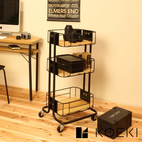黑色三層置物推車，採用金屬框架搭配木質層板，底部有四個萬向輪，推車上擺放有相機鏡頭、黑色收納盒及紙箱。