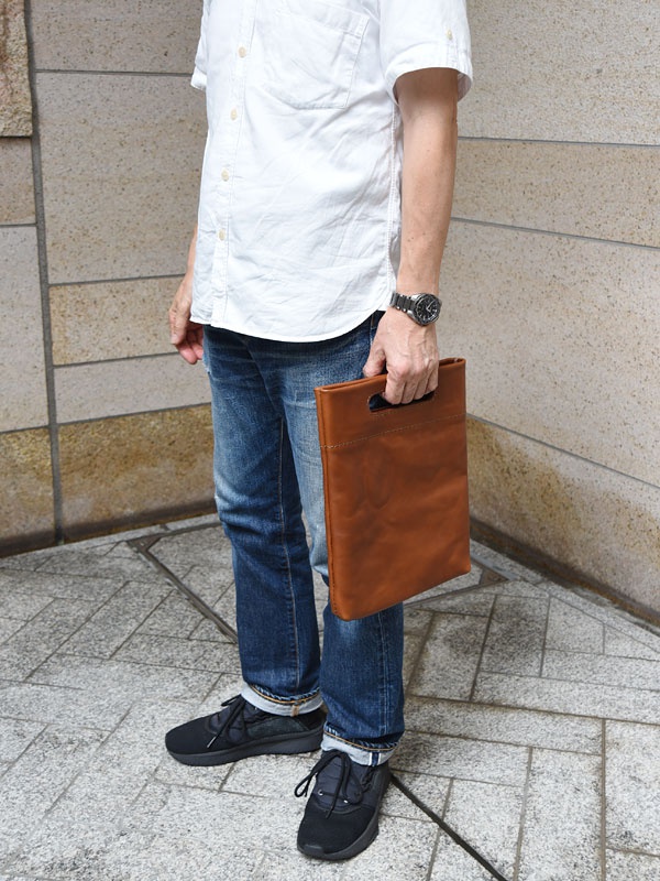 一名男子手持一個棕色皮革直式收納袋，袋子上有一個長方形開口手把。