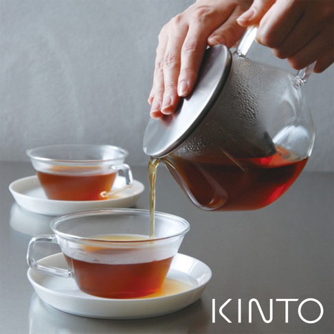 透明玻璃茶壺，內含茶葉，正將琥珀色熱茶倒入另一個透明玻璃茶杯中。茶壺帶有金屬濾網和手把。