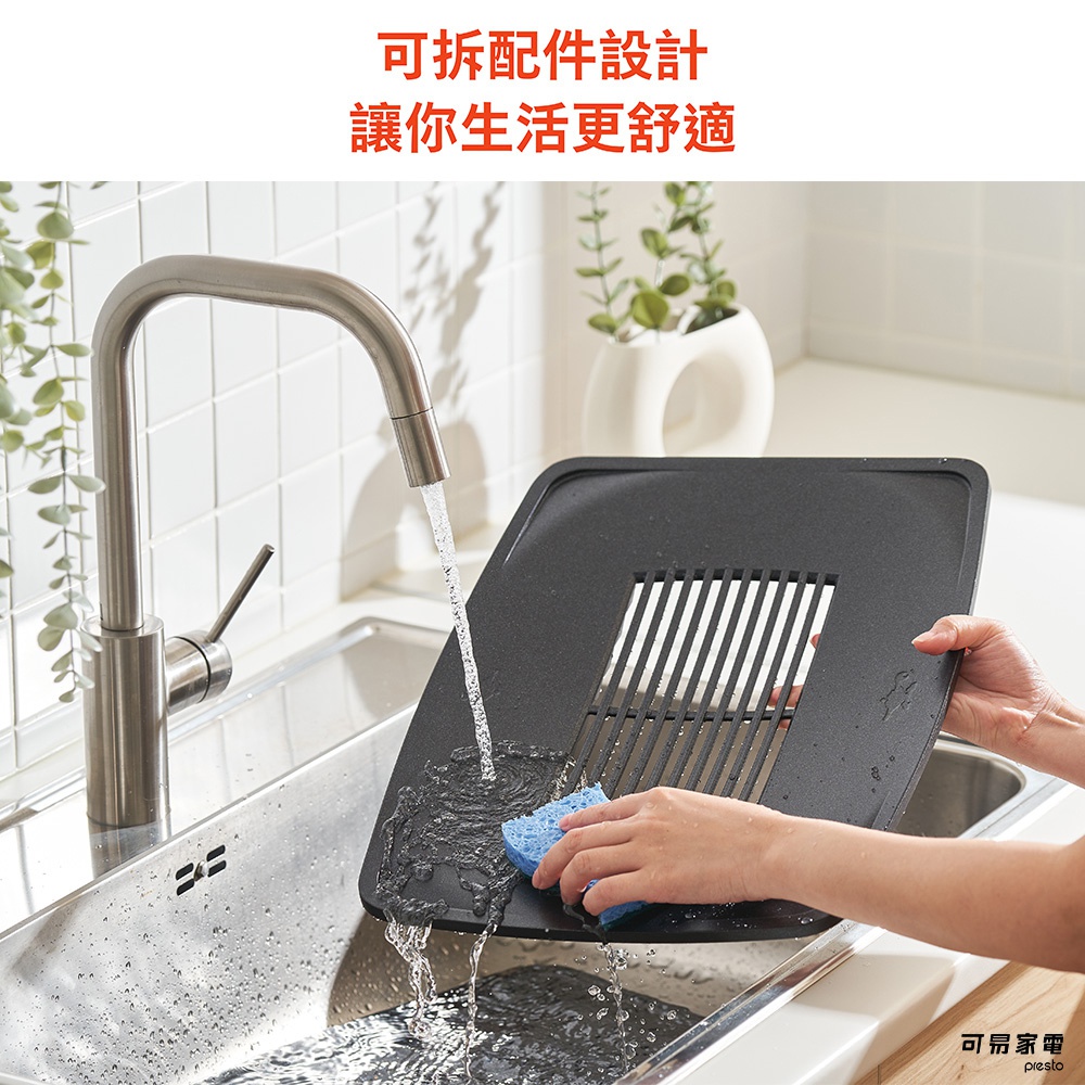 黑色方形烤盤，表面有條狀紋理，一隻手拿著藍色海綿正在擦拭，水從水龍頭流下，沖洗烤盤。