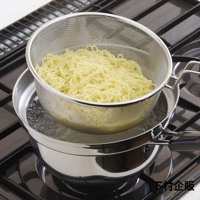 不鏽鋼防溢汆燙鍋，附帶一個細網篩，內盛煮熟的黃色麵條，鍋中裝有冒泡的沸水，置於瓦斯爐上。