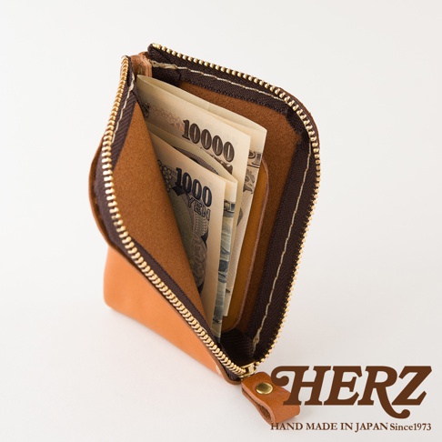 一個採用淺褐色皮革製成的日本HERZ職人手作迷你錢包，錢包內部設有夾層並放入了日幣紙鈔，側邊拉鍊設計精緻，整體呈現出高品質的手工質感與簡約的視角風格。