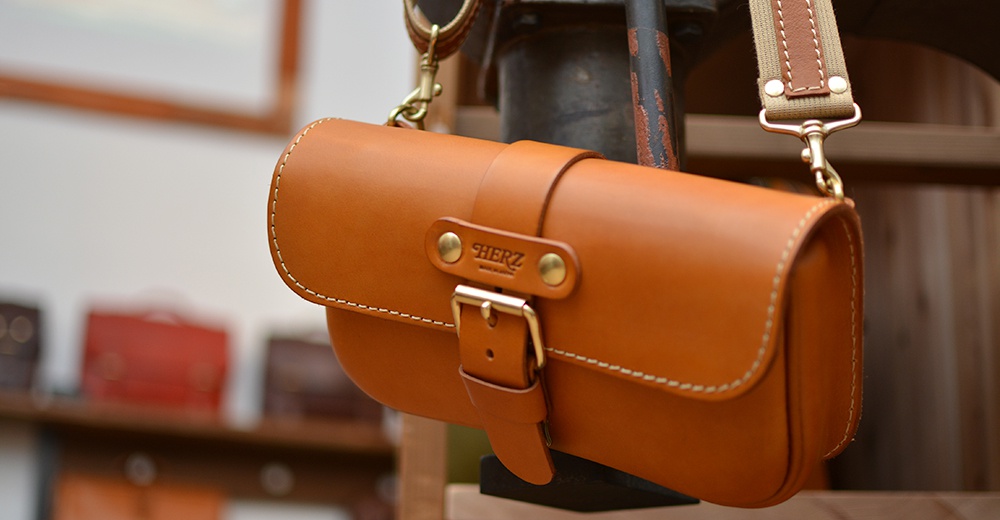 職人手作皮革單扣經典斜背包，呈現溫暖的琥珀棕色，包身以精緻的白色縫線勾勒，搭配金屬釦環與品牌標誌，風格簡約而質感。