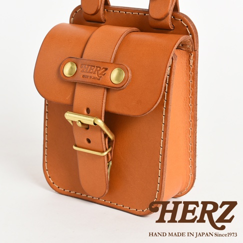 職人手作的經典皮革腰袋，採用光滑的棕色皮革製成，配有黃銅色鈕扣和皮帶扣。袋蓋上有「HERZ」品牌標誌，並以手工縫線裝飾。