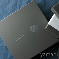 Tsudoi 赤飴釉美濃燒圓盤禮盒，深灰色紙盒，印有白色「Tsudoi」字樣及銀色螺旋花紋。部分露出另一黑色禮盒，其中包裝著淡綠色緞帶。